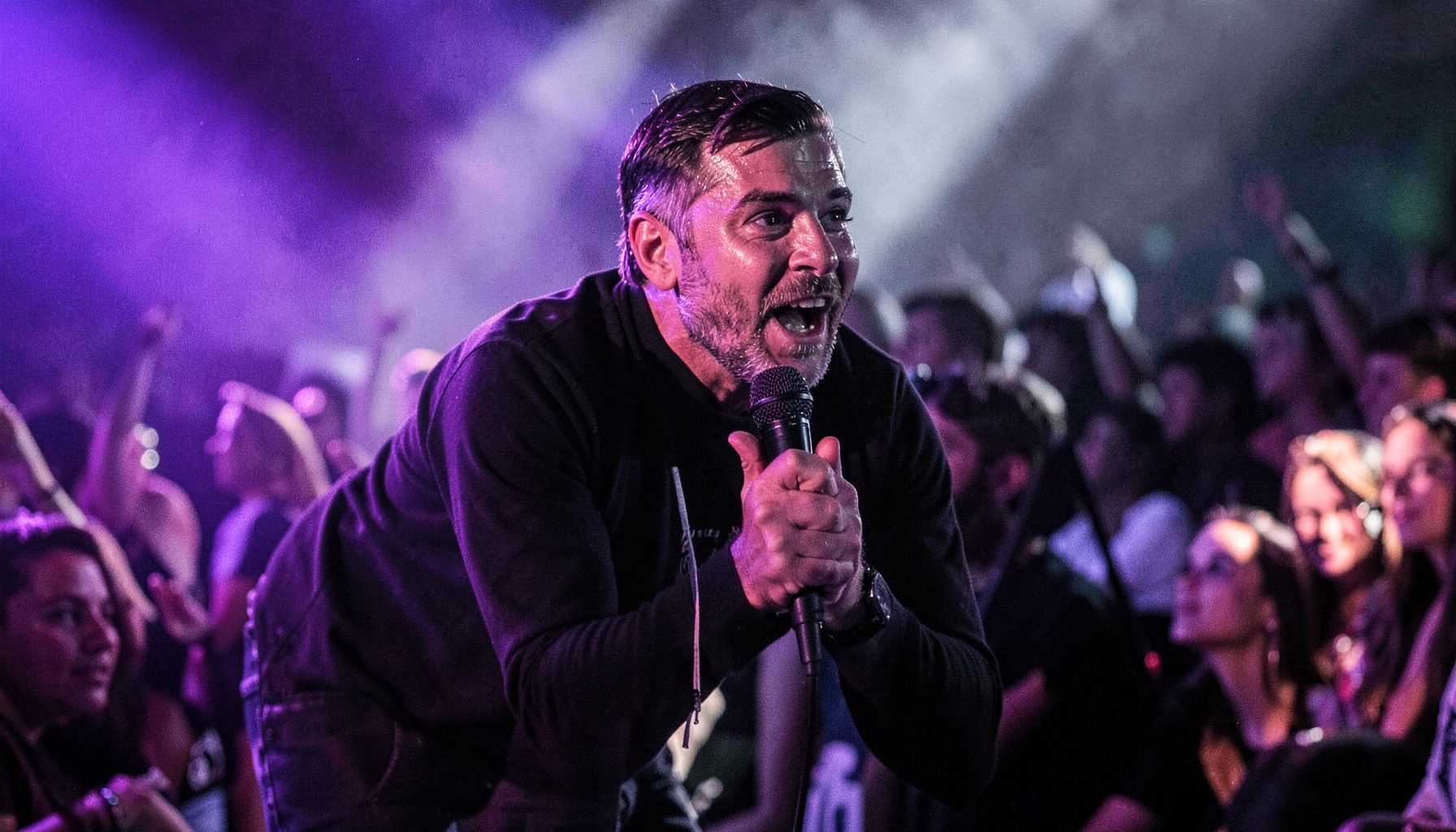 Singer leaning into the audience, microphone in hand, dramatic side lighting in purple and white