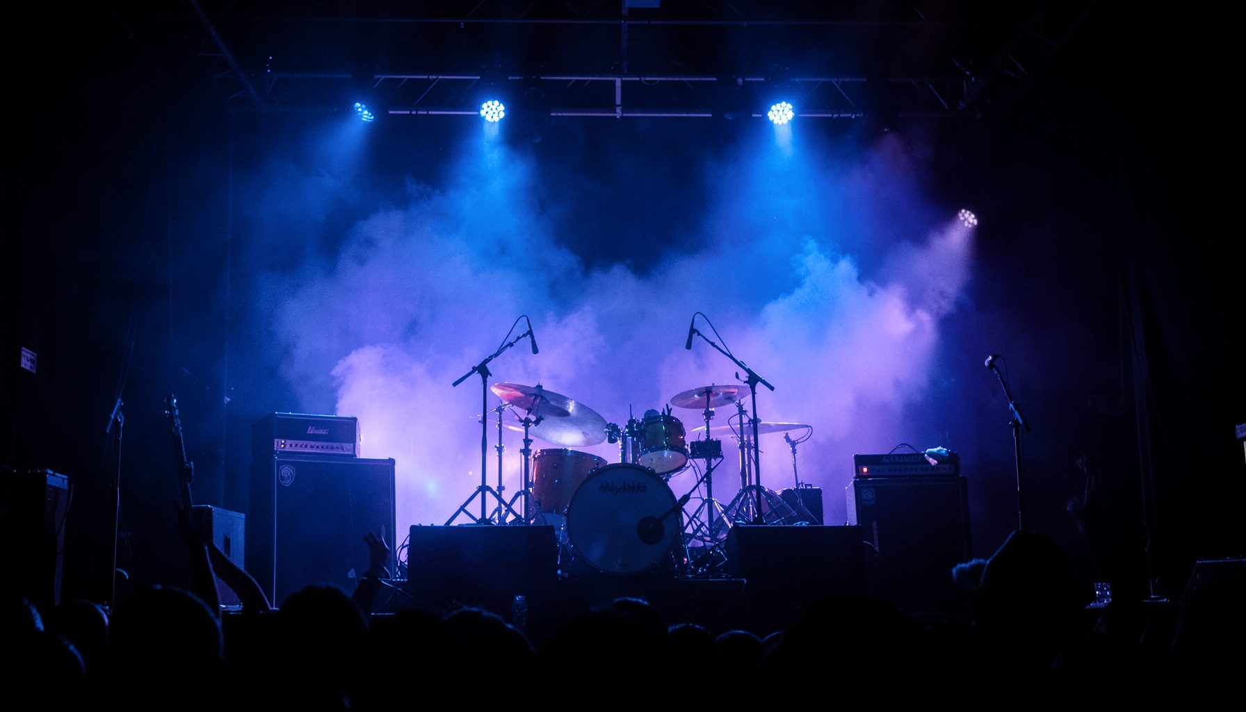 Dark concert stage with dramatic blue and purple lighting, amplifiers and drum kit silhouetted against the haze