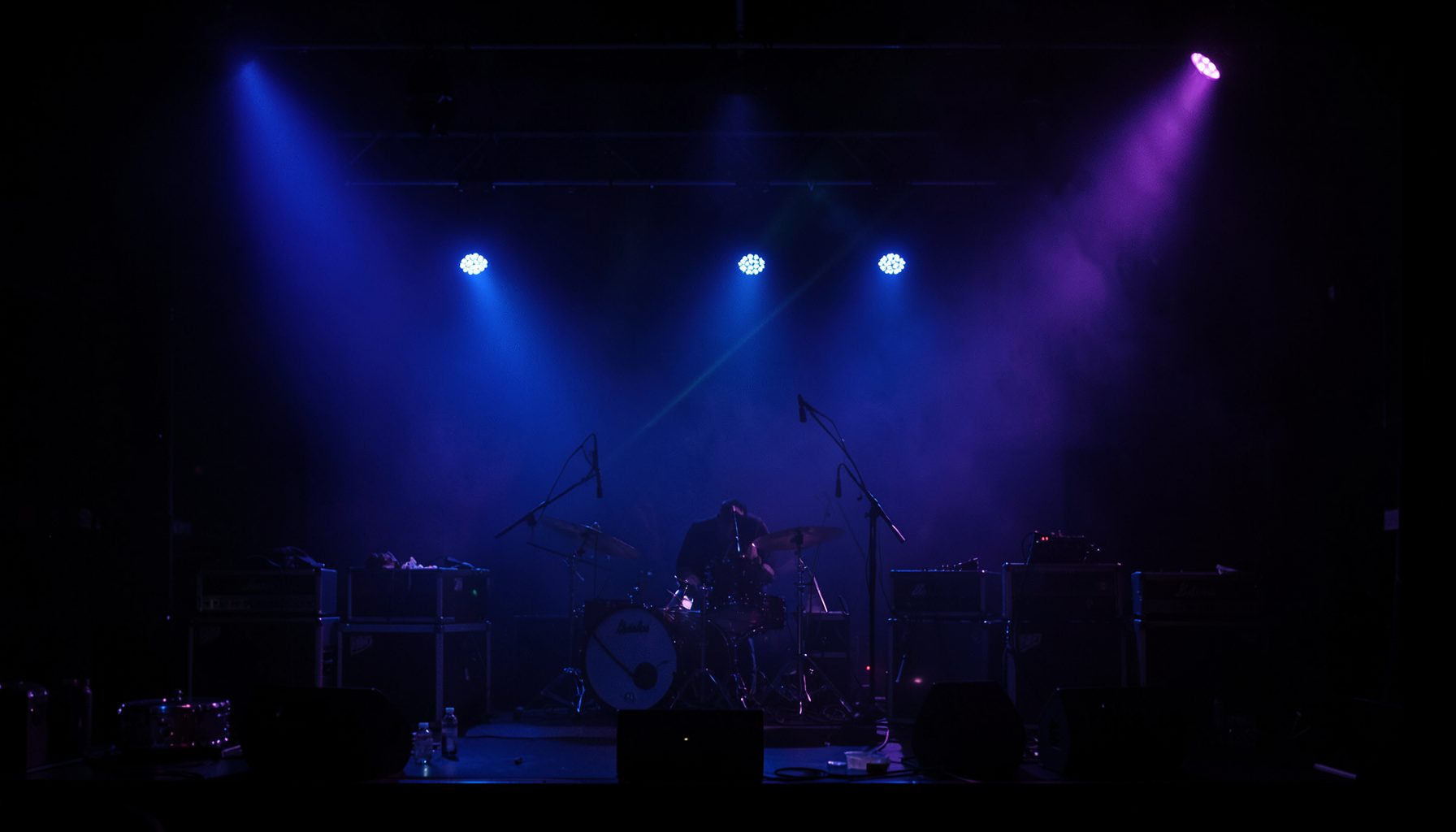 Dark concert stage lit by deep blue and purple spotlights, with a hazy atmosphere and silhouetted amplifiers