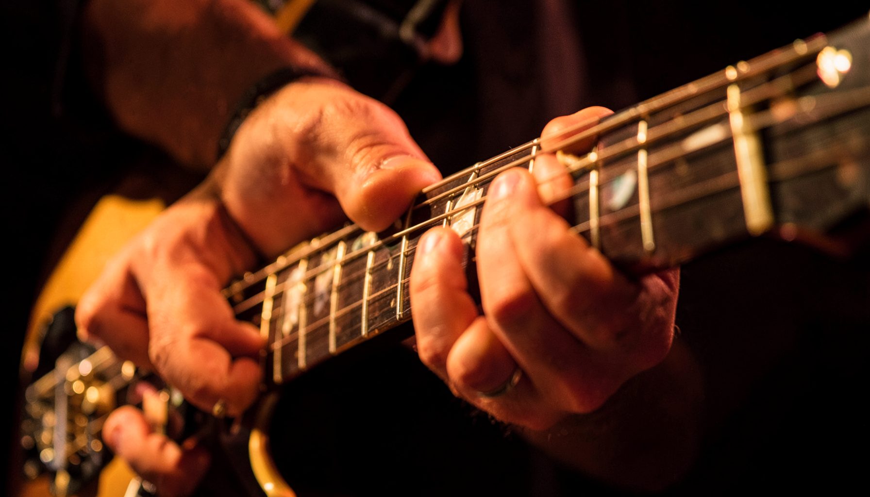 Close-up of a guitarist's hands on a guitar neck under warm amber stage lights, strings and frets in sharp focus