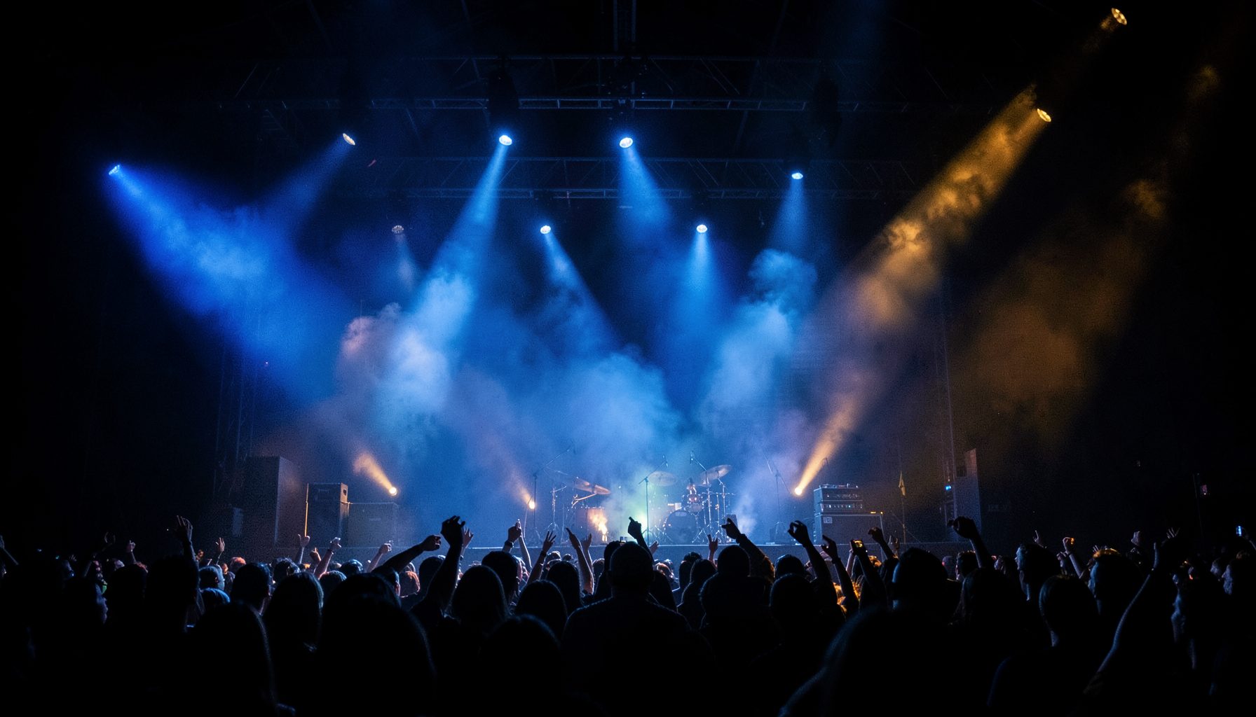 Dark stage with dramatic blue and amber spotlights cutting through haze, a rock concert atmosphere