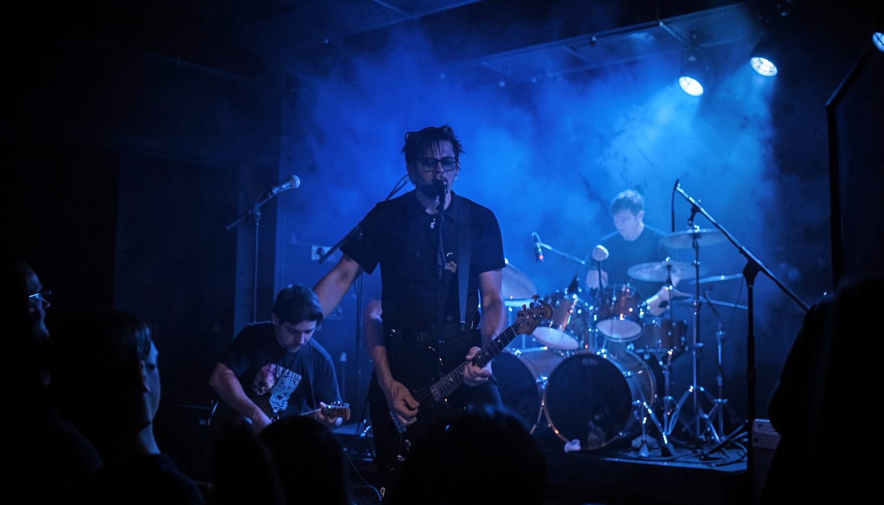 Rock band performing in a dimly lit club, blue stage wash, drummer visible in the background
