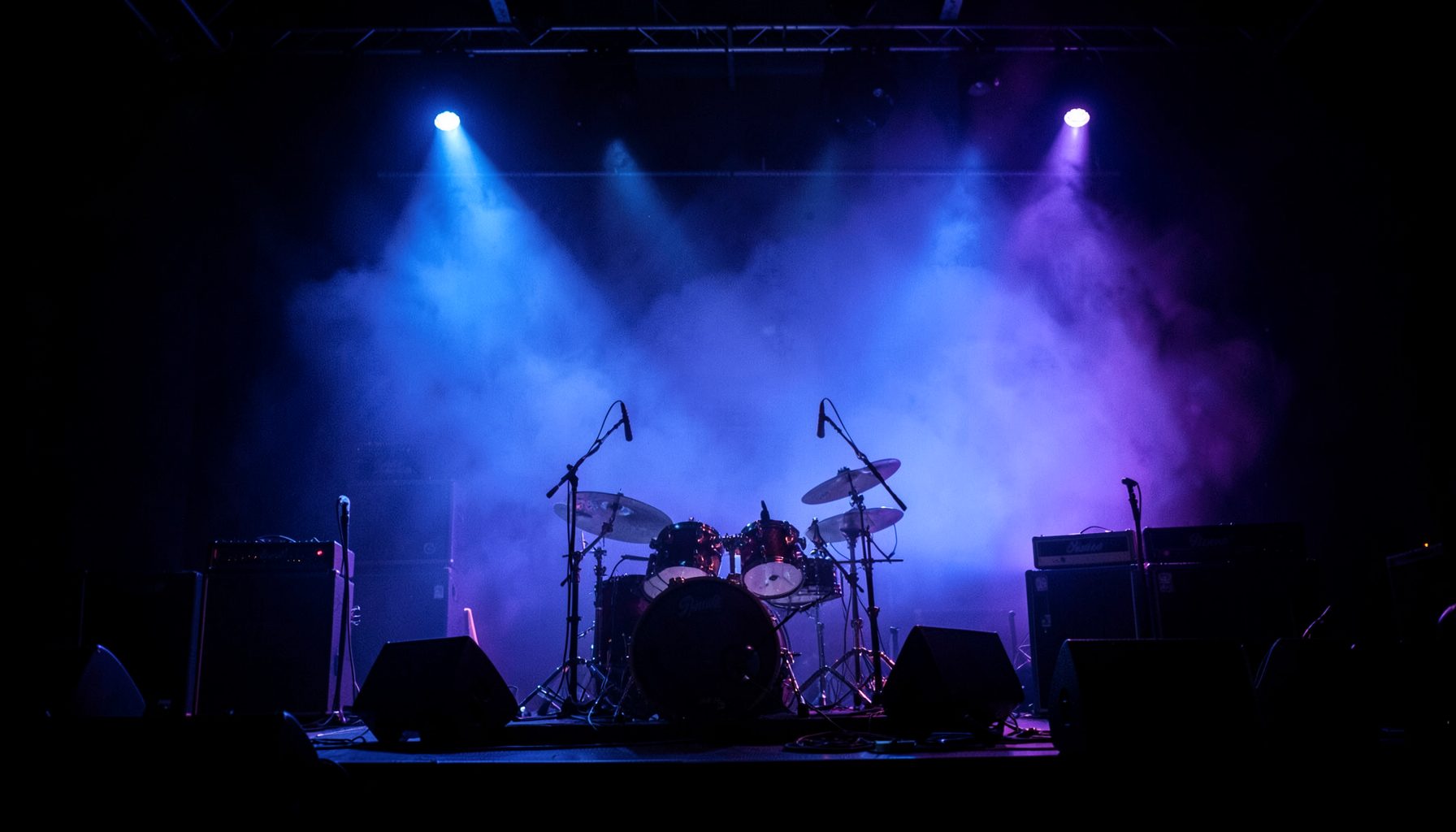 Dark concert stage with dramatic blue and purple lighting, amplifiers and drum kit silhouetted against a hazy backdrop